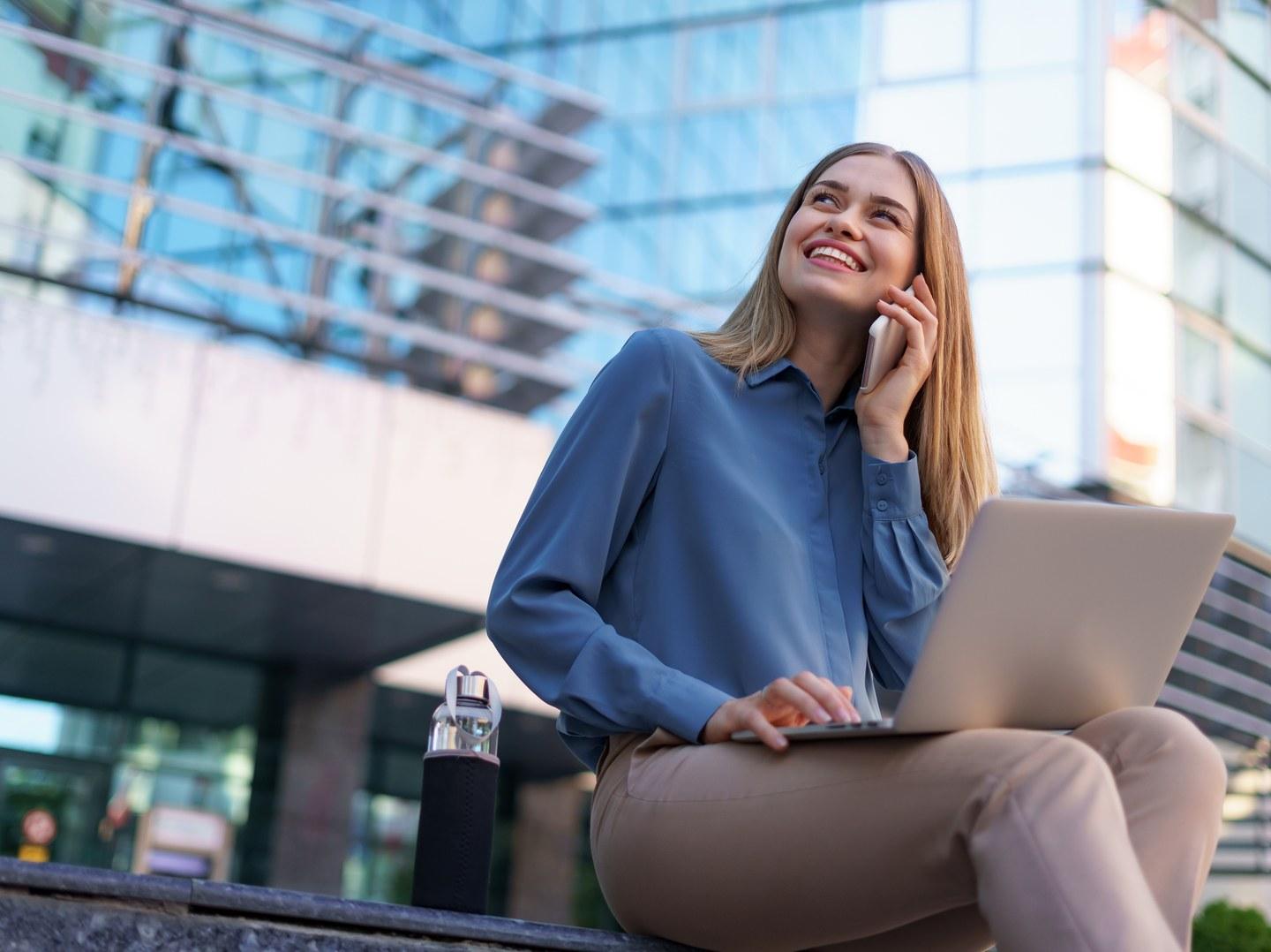 Mulher ao telefone usando o notebook em frente a um prédio comercial